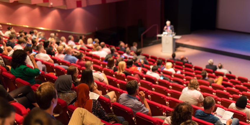 Photograph of an auditorium, with an audience watching a presentation by a speaker at a lectern