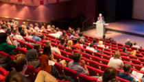 Photograph of an auditorium, with an audience watching a presentation by a speaker at a lectern